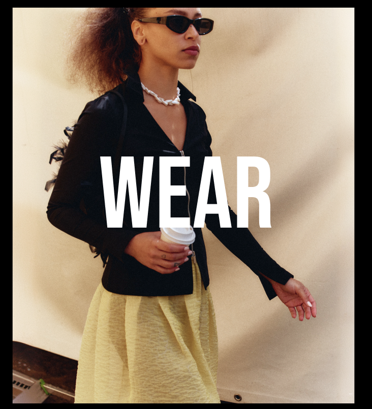 A woman wearing black sunglasses, a black jacket, and a light yellow skirt walks while holding a coffee cup. Large white text reading 'WEAR' is overlaid across the center.