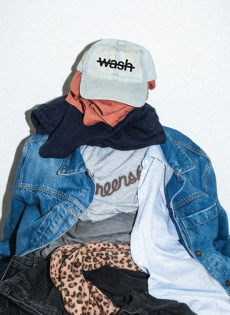 A styled pile of clothing on a wood floor against a white wall, featuring a light blue cap with 'week' embroidered on it, a blue denim jacket, a white t-shirt, a navy sweatshirt, a gray tee, leopard-print pants, and black jeans.